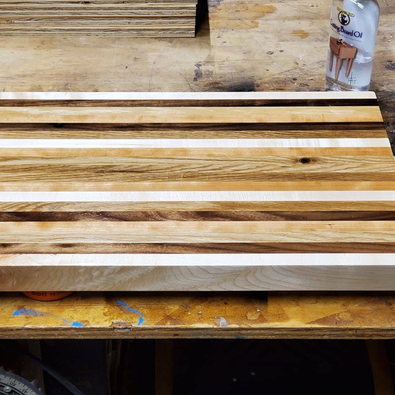 May include: A large, rectangular cutting board made of multiple wood types. The board has a light brown, white, and dark brown striped pattern. The board is sitting on a wooden surface.