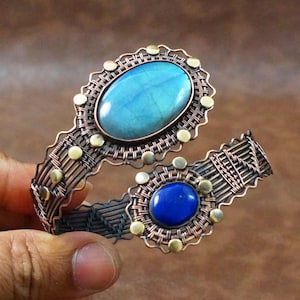 May include: A wire-wrapped cuff bracelet crafted from dark copper wire, featuring a large oval blue labradorite stone and a smaller round deep blue lapis lazuli stone. The design includes decorative brass-colored metal studs along the edges.