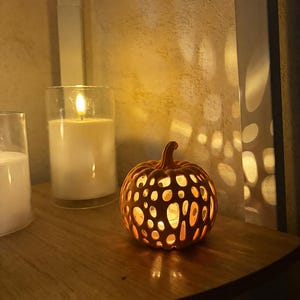 Op de afbeelding: Een kleine oranje pompoenvormige lantaarn met een hol, organisch, webachtig uitgesneden patroon. De pompoen wordt van binnenuit verlicht, werpt zacht licht door de openingen en staat naast een hoge glazen kaarsenhouder.
