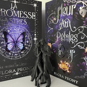 Puede incluir: Dos libros con cubiertas oscuras y una figura negra de una pareja abrazándose. Los libros tienen detalles morados y plateados, con los títulos "La Promesse Des Astres" y "Fleur Aux Pétales D'Or". La figura está delante de los libros.