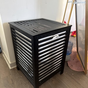 May include: A black wooden laundry hamper with a slatted design. The hamper has a square shape with a flat top and four legs. The interior is lined with a white fabric bag.