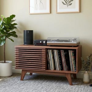 May include: A mid-century modern style wooden record player console with a slatted door, holding vinyl records. It features a turntable, amplifier, and a black speaker. A potted plant and framed art are in the background.