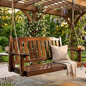 May include: A wooden porch swing with a slatted back and armrests hangs from chains attached to a wooden pergola. A beige pillow and blanket rest on the swing. The background features a garden with flowers and greenery.