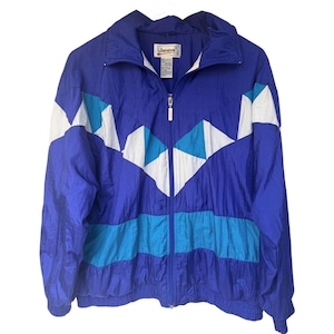 May include: A royal blue windbreaker jacket featuring a high collar, a full-length front zipper, and a geometric chevron pattern across the chest in white and turquoise. The jacket includes a horizontal turquoise stripe and a Janeve brand label.