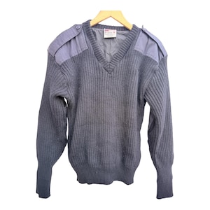 Może przedstawiać: Szary sweter z dekoltem w serek i naszywkami na ramionach. Sweter ma prążkowaną dzianinę i długie rękawy. Naszywki na ramionach są w jaśniejszym odcieniu szarości. Sweter wisi na drewnianym wieszaku.