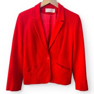 May include: A vibrant red blazer featuring a single-button closure, notched lapels, and two front welt pockets. The jacket is fully lined in a matching red fabric and includes a white interior brand label at the neckline, hanging on a wooden hanger.