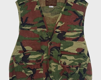 Op de afbeelding: Een mouwloos vest met een camouflagepatroon in tinten groen, bruin en zwart. Het kledingstuk heeft vier voorzakken met knoopsluiting en een label bij de nek met de tekst Caliber Sportsman Apparel maat L.