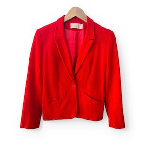 Op de afbeelding: Een felrode blazer met een klassiek ontwerp, met een revers, twee voorzakken en een sluiting met één knoop. De blazer hangt aan een houten kleerhanger tegen een witte achtergrond. De binnenvoering van de jas is ook rood.
