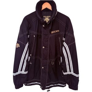 May include: A black Descente jacket featuring a high collar, a front zipper closure with metal snap buttons, and white striped detailing on the sleeves. The jacket includes multiple zippered pockets, a D-ring attachment, and the Descente logo.
