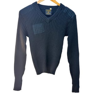 May include: A dark navy blue V-neck sweater featuring a thick ribbed knit texture, long sleeves, and reinforced fabric patches on the shoulders and chest. A rectangular label inside the neckline displays the brand name Citadel in gold text.