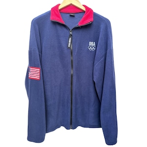 Op de afbeelding: Een marineblauw fleece jack met rits en een felroze binnenkraag. Op de voorzijde staat in witte letters USA boven het logo met de olympische ringen. De zwarte ritstrekker is voorzien van de tekst Team USA en een Amerikaanse vlag op de mouw.