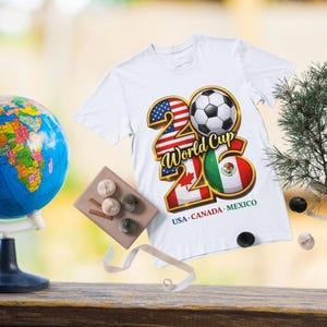 Pode incluir: Camiseta branca com um gr&aacute;fico colorido para a Copa do Mundo de 2026. O design inclui uma bola de futebol, os n&uacute;meros 20 e 26 e as bandeiras dos EUA, Canad&aacute; e M&eacute;xico. O texto "World Cup" tamb&eacute;m est&aacute; presente.