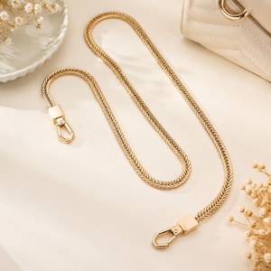May include: A gold-toned metal chain strap for a handbag, featuring a flat herringbone link design. The strap includes a polished gold-coloured lobster clasp at each end, designed for easy attachment to a bag's hardware rings or loops.
