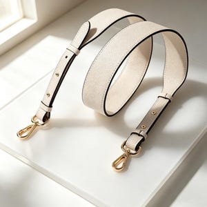 May include: Cream-colored handbag strap with black edging and gold-tone hardware. The strap is made of a textured material and features adjustable length with metal clasps for attachment. The design is simple and elegant, suitable for various bag styles.