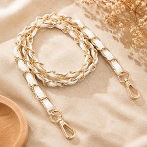 May include: A replacement handbag strap featuring a gold-tone metal chain interwoven with a white leather-like material. The strap is coiled in a circular shape and includes two gold-tone lobster claw clasps at each end for secure attachment.
