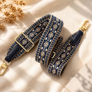 May include: A patterned bag strap in navy blue, gold, and white. The strap has a geometric design and gold-coloured metal hardware, including a buckle and clasps. The strap is made of woven fabric with leather detailing.