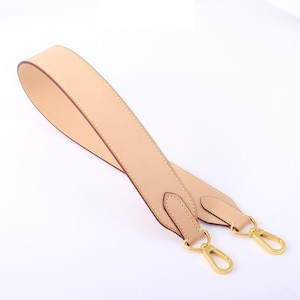 May include: A light beige leather bag strap with gold-colored metal clasps. The strap is stitched with a contrasting thread and has a smooth, finished appearance. The clasps are attached to the strap with small leather tabs.