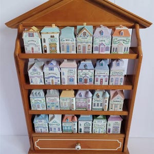 Pode incluir: Um porta-especiarias de madeira com design em forma de casa, com 24 potes de especiarias de cerâmica. Cada pote é pintado para se assemelhar a uma casa em miniatura, com rótulos como "Manjericão" e "Canela". O porta-especiarias tem uma gaveta com um botão branco.