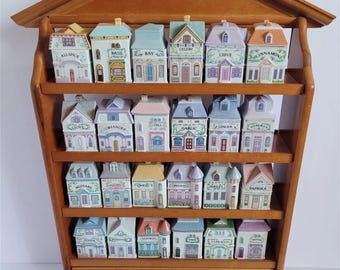 May include: A wooden spice rack with a house-shaped design, holding 24 ceramic spice jars. Each jar is painted to resemble a miniature house, with labels like "Basil" and "Cinnamon." The rack has a drawer with a white knob and is ideal for kitchen decor.