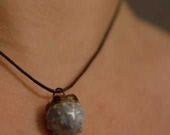 May include: A small, spherical ceramic pendant with a glossy light blue finish and raised cream-coloured dots, suspended from a thin, dark brown cord. The pendant features a dark, textured top section and a smooth, rounded base.