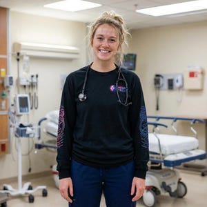 May include: A smiling person wearing a black long-sleeve shirt with the text "NEURO TECH" and a stethoscope around their neck. The shirt has a logo on the chest and arm. The person is standing in a medical setting.