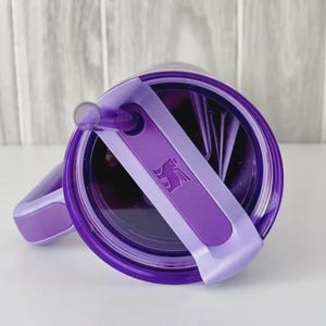 May include: A purple tumbler with a matching lid and straw. The lid features a sliding closure with a logo. The tumbler has a handle and is designed for cold beverages.