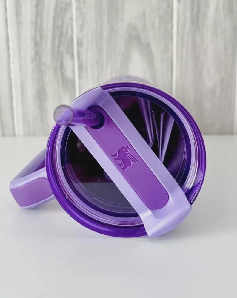 May include: A purple tumbler with a matching lid and straw. The lid features a sliding closure with a logo. The tumbler has a handle and is designed for cold beverages.