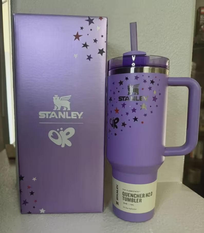 May include: A purple Stanley Quencher H2.0 tumbler with a handle and straw, next to a matching box. Both the tumbler and box feature a star pattern and the Stanley logo. The tumbler has a capacity of 40 oz.