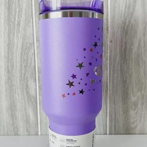 May include: A purple insulated tumbler with a clear lid and straw. The tumbler is decorated with star cutouts in various colors. A white label with product information is wrapped around the base.