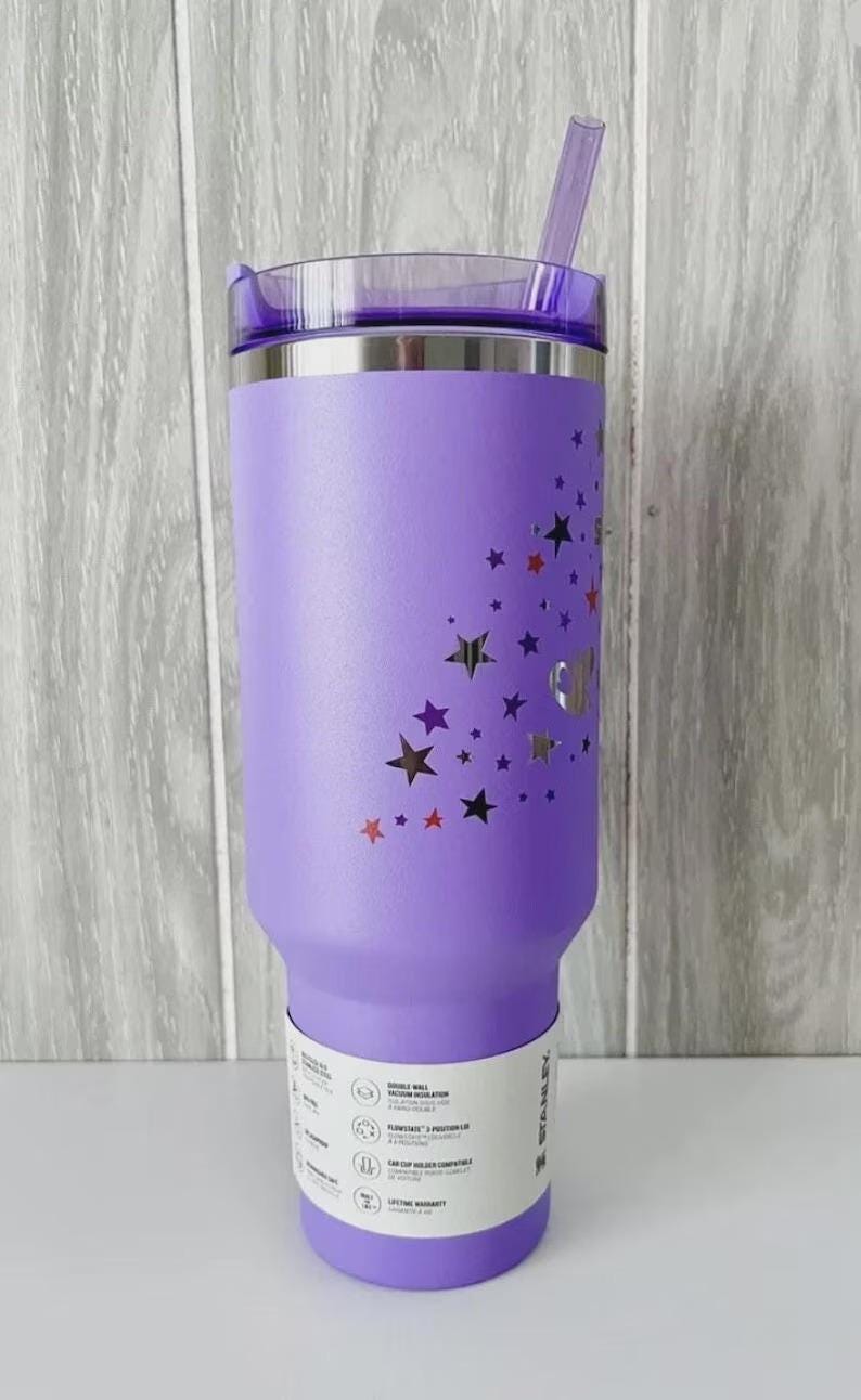 May include: A purple insulated tumbler with a clear lid and straw. The tumbler is decorated with star cutouts in various colors. A white label with product information is wrapped around the base.
