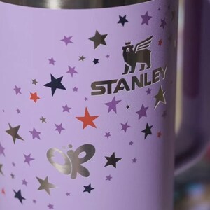 May include: A lavender Stanley mug with a silver rim and handle. The mug is decorated with gold and silver stars, a butterfly design, and the Stanley logo. The logo features a winged animal wearing a crown.