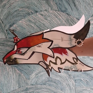 Può includere: Una testa di drago di carta fatta a mano e stilizzata in rosso, bianco e nero. Il drago presenta un design dettagliato con un orecchino a forma di ingranaggio e un corno bianco. L'opera d'arte è ambientata su uno sfondo blu strutturato.