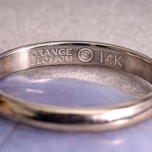 May include: A close-up of a gold ring with the text "ORANGE BLOSSOM" and "14K" engraved on the band. The ring is set against a light blue surface. The ring is a classic jewelry piece.