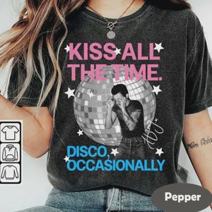 May include: Dark gray t-shirt with the text "KISS ALL THE TIME" in pink and "DISCO, OCCASIONALLY" in blue. A black and white image of a person is in front of a disco ball. White stars are scattered around the design.