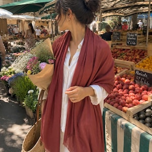 May include: A solid terracotta-colored rectangular scarf with a fringed hem, draped over a white button-down shirt. The scarf features a lightweight, woven texture and hangs loosely around the shoulders, providing a simple and functional accessory.
