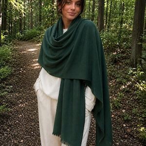 May include: A dark forest green rectangular scarf with a soft, fine-knit texture and delicate fringe detailing along the edges. The scarf is draped loosely around the shoulders and neck, layered over a cream-colored long-sleeve top and matching pants.