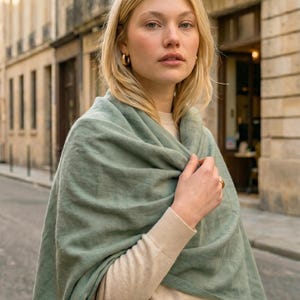 Op de afbeelding: Een vrouw draagt een saliegroene sjaal over een beige trui en houdt een taupe handtas vast. Ze heeft blond haar en gouden oorringen. De achtergrond toont een Europese straat met stenen gebouwen.
