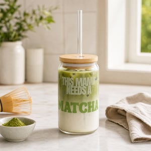 Op de afbeelding: Een doorzichtig glas met een bamboe deksel en een glazen rietje, gevuld met een gelaagde matcha latte. Het glas is voorzien van de witte tekst "THIS MAMA NEEDS A MATCHA" op de voorzijde, met een verloop van groene matcha en witte melk.