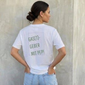 Op de afbeelding: Wit T-shirt met de tekst "GAS(T)-GEBER MIT PEP!" in groene letters in een groene rechthoek. Het shirt heeft korte mouwen en wordt gedragen door een persoon met de handen in de zakken. De persoon draagt een lichtblauwe jeans.