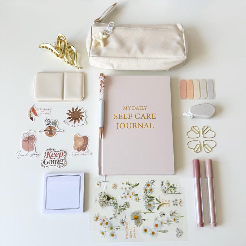 K&ouml;nnte beinhalten: Eine Auswahl an Selbstpflegeartikeln, darunter ein Notizbuch mit dem Text "My Daily Self Care Journal", Aufkleber, Stifte, ein Federm&auml;ppchen und eine Haarspange. Die Artikel sind auf einer wei&szlig;en Oberfl&auml;che angeordnet. Die Farbpalette ist sanft, mit Rosa-, Creme- und Goldakzenten.