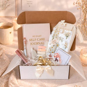 K&ouml;nnte beinhalten: Eine Selbstpflege-Geschenkbox gef&uuml;llt mit einem Tagebuch, Stiften, Aufklebern und anderen Artikeln. Die Box ist ge&ouml;ffnet und zeigt eine Vielzahl von Produkten, darunter ein Tagebuch mit dem Text "MY DAILY SELF CARE JOURNAL". Die Box ist mit einer cremefarbenen Schleife gebunden.