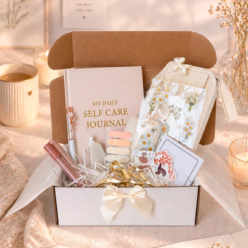K&ouml;nnte beinhalten: Eine Selbstpflege-Geschenkbox gef&uuml;llt mit einem Tagebuch, Stiften, Aufklebern und anderen Artikeln. Die Box ist ge&ouml;ffnet und zeigt eine Vielzahl von Produkten, darunter ein Tagebuch mit dem Text "MY DAILY SELF CARE JOURNAL". Die Box ist mit einer cremefarbenen Schleife gebunden.