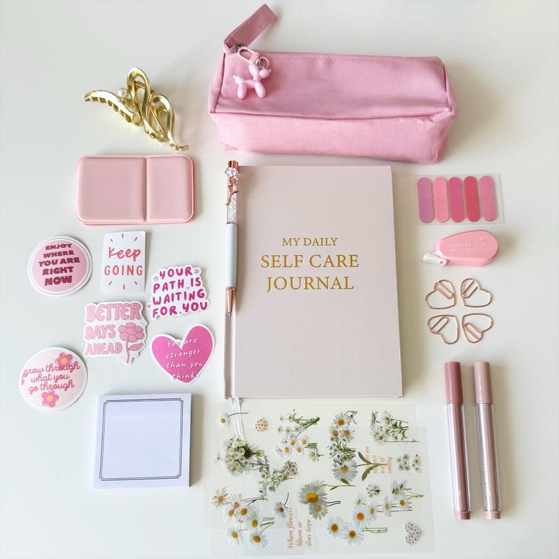K&ouml;nnte beinhalten: Eine Sammlung von Selbstpflegeartikeln in Rosa- und Goldt&ouml;nen, darunter ein Notizbuch, ein Stift, Aufkleber und ein Federm&auml;ppchen. Auf dem Notizbuch steht "MY DAILY SELF CARE JOURNAL". Dekorative Elemente sind eine goldene Haarspange und herzf&ouml;rmige B&uuml;roklammern.