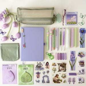 May include: A collection of stationery items including a lavender notebook, a translucent green pencil case, a small green pouch, various pens, sticky notes, and decorative stickers featuring floral and landscape illustrations in shades of purple and green.
