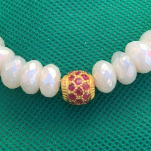 May include: A beaded necklace featuring a series of faceted, iridescent white beads arranged in a row. A single, spherical gold-toned bead encrusted with small, circular red stones is positioned centrally among the white beads.