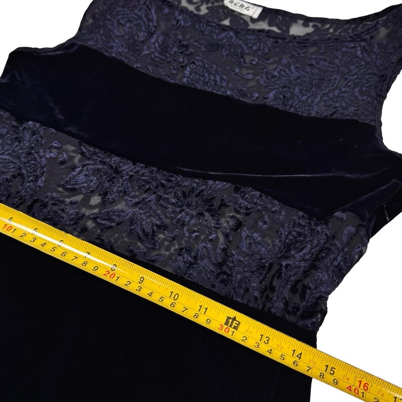 May include: A dark blue sleeveless dress featuring a velvet bodice and a sheer, floral-patterned overlay. A thick velvet band accents the midsection. The dress is being measured with a yellow tape measure.