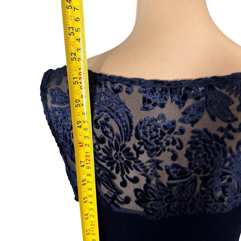 May include: A navy blue dress with a sheer overlay featuring a floral velvet pattern. The dress has a boat neckline and is being measured with a yellow measuring tape. The dress is approximately 109 to 137 cm in length.
