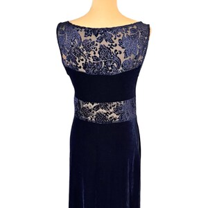 May include: A navy blue velvet evening gown. The dress features a sheer, floral-patterned overlay on the upper back and waist. The dress has a scoop neckline and a fitted bodice that flows into a floor-length skirt.