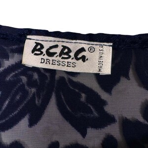 May include: Close-up of a navy blue dress with a floral pattern. The label reads "B.C.B.G. DRESSES" and "MADE IN U.S.A." in white text. The dress has a sheer overlay.