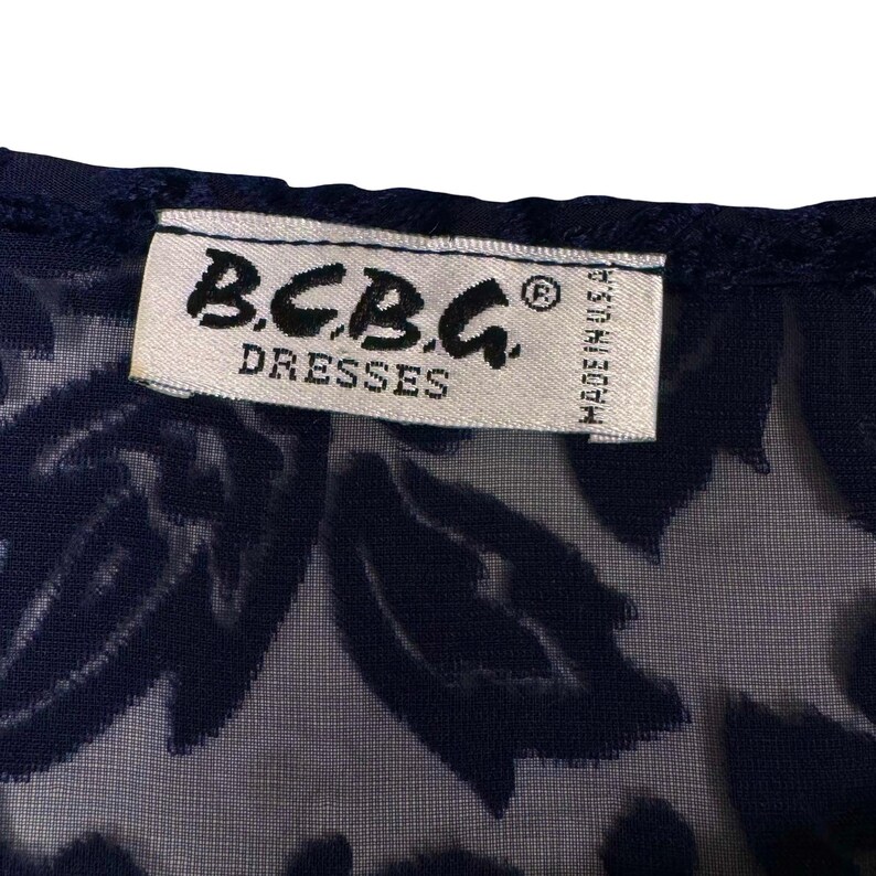 May include: Close-up of a navy blue dress with a floral pattern. The label reads "B.C.B.G. DRESSES" and "MADE IN U.S.A." in white text. The dress has a sheer overlay.